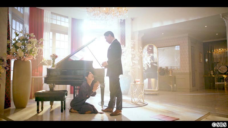 ピアノの前でしゃがみ込む女性　その右に立つ男性