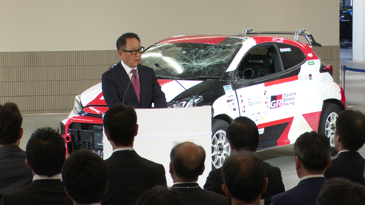 男性が登壇している。後ろには大破した車が展示されている。